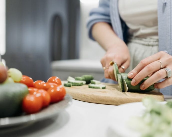 Close-up of a woman slicing cucumbers on a wooden board surrounded by fresh vegetables.