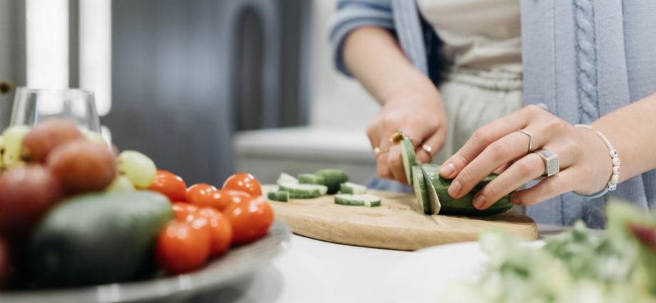 Close-up of a woman slicing cucumbers on a wooden board surrounded by fresh vegetables.